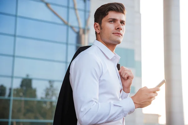 Pensive young businessman holding mobile phone with jacket over shoulder — Stock Photo, Image