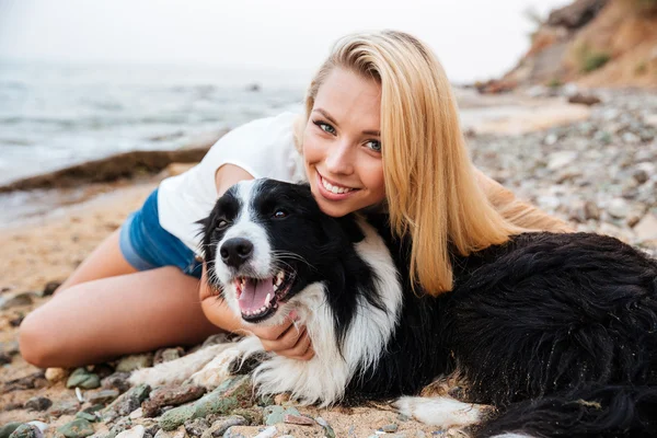 Cheerful woman sitting and hugging her dog on the beach — Stock Photo, Image