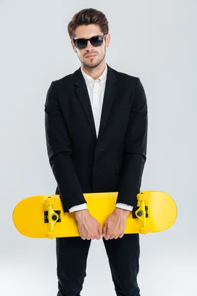 Attraktiver junger Geschäftsmann im schwarzen Anzug mit gelbem Skateboard — Stockfoto