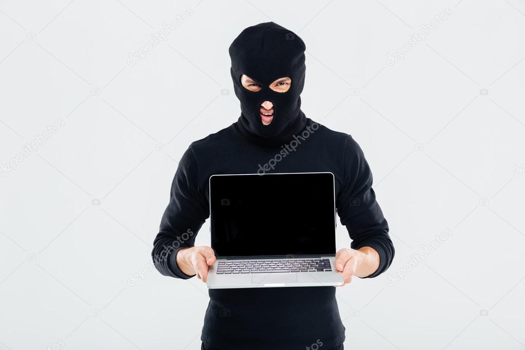 Criminal man in balaclava holding blank screen laptop