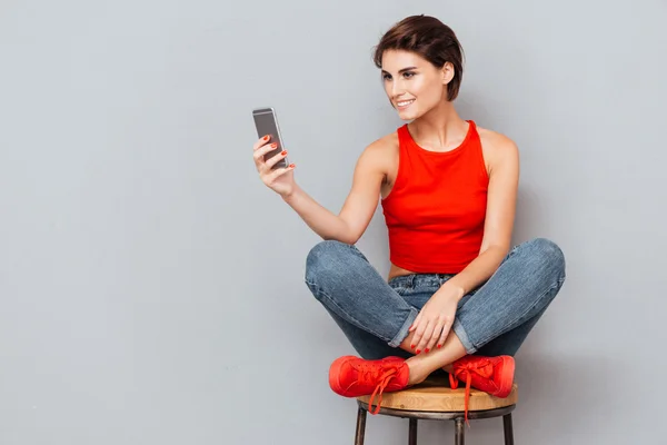 Lächelnde brünette Frau, die auf dem Stuhl sitzt und das Smartphone benutzt — Stockfoto