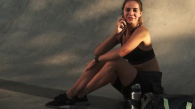 Gülümseyen genç bir kadın dışarıda spor salonunda akıllı telefonuyla konuşuyor.