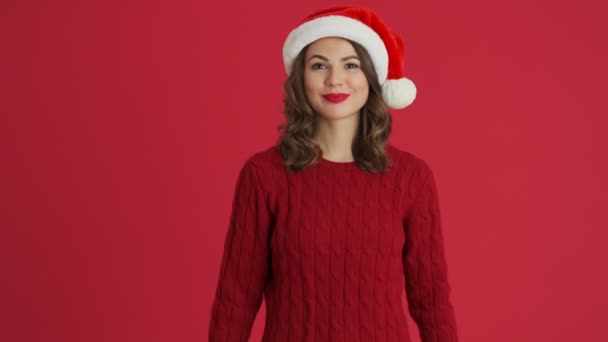 暖かい赤いセーターとクリスマスの帽子を身に着けている笑顔の女性は 赤い背景の上に隔離された立ってこんにちはジェスチャーをしています — ストック動画