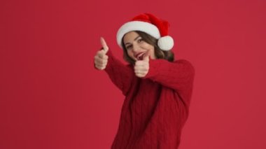Sıcak kırmızı bir kazak ve Noel şapkası giyen neşeli bir kadın iki eli kırmızı arka planda izole bir şekilde baş parmak hareketi yapıyor.