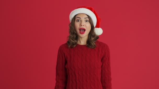 暖かい赤いセーターとクリスマスの帽子を身に着けている驚きの女性は喜んでいます彼女の手を閉じ込めながら彼女の手は赤い背景の上に隔離 — ストック動画