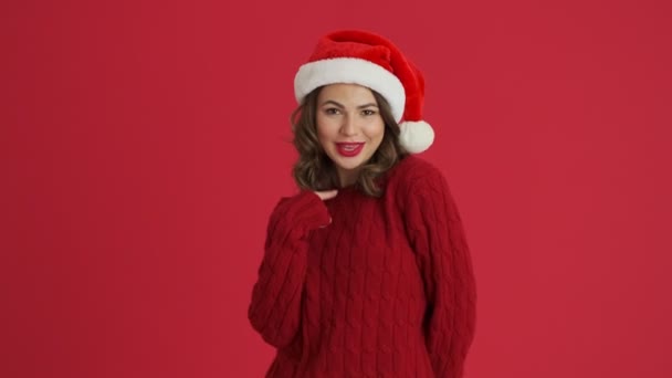 暖かい赤いセーターとクリスマスの帽子を身に着けている魅力的な女性は心を示しており 赤い背景の上に隔離された立っている視聴者を指しています — ストック動画