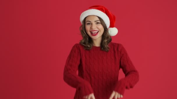 暖かい赤いセーターとクリスマスの帽子を身に着けている笑顔の女性は 赤い背景の上に隔離された空中に大きなハートサインを描いています — ストック動画