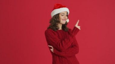 Sıcak kırmızı süveter ve Noel şapkası giyen kendine güvenen bir kadın kırmızı arka planda izole bir şekilde duran boş alandaki noktaları işaret ediyor.