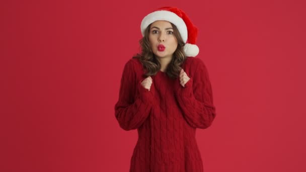暖かい赤いセーターとクリスマスの帽子を身に着けている美しい女性は寒さを感じています立ち赤の背景に隔離 — ストック動画