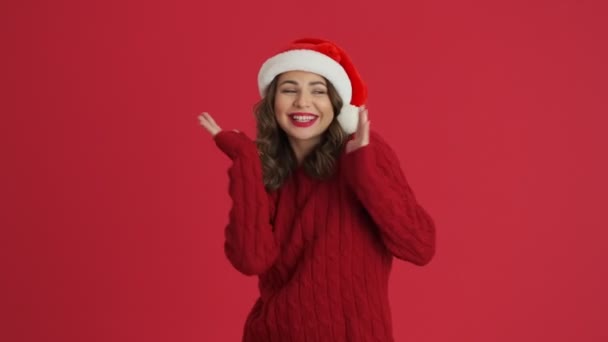 暖かい赤いセーターとクリスマスの帽子を身に着けている幸せな女性は赤い背景に孤立して踊っています — ストック動画