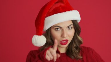 Sıcak kırmızı bir kazak ve Noel şapkası giyen pozitif bir kadının yakından görüntüsü, kalp şarkısını gösteriyor ve kırmızı arka planda izleyiciyi işaret ediyor.