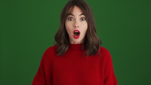 緑の背景に隔離されたカメラを見ながら赤いセーターのショックを受けた女性が口を開けている — ストック動画