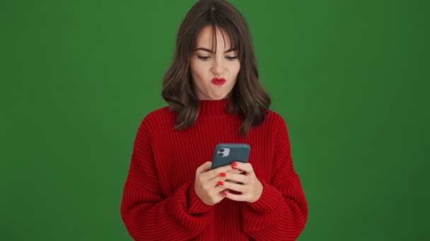 赤いセーターの若い女性は 緑の背景に隔離されたスマートフォンを使用しています — ストック動画