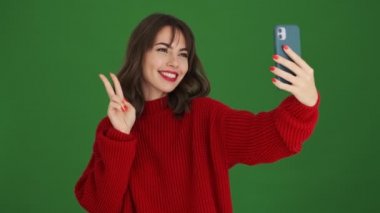 Kırmızı kazaklı güzel bir kadın yeşil arka planda tek başına duran akıllı telefonuyla selfie çekiyor.