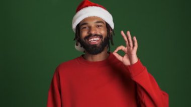 Noel Baba şapkalı genç, iyimser Afrikalı adam yeşil duvar arkasında izole edilmiş.