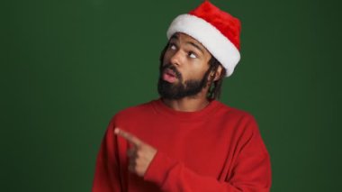 Noel Baba şapkalı genç, iyimser Afrikalı adam yeşil duvar arkaplanını işaret ediyor.