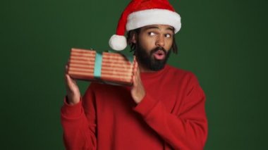 Noel Baba şapkalı genç, pozitif Afrikalı bir adam yeşil duvardan izole edilmiş, elinde hediye kutusu var.