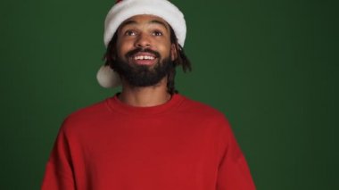 Mutlu, mutlu, Afrikalı, Noel Baba şapkalı genç adam yeşil duvarın arkasında elinde hediye kutusuyla izole edilmiş.