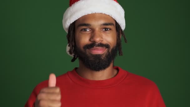 戴着圣诞圣诞圣诞礼帽的年轻快乐的非洲男人被隔离在绿墙的背景之外 露出了竖起大拇指的姿势 — 图库视频影像