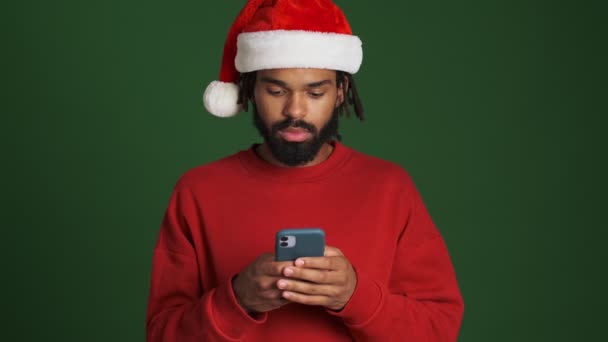 若いです悲しいアフリカ人男性でクリスマスサンタ帽子隔離使用してスマートフォン以上緑の壁の背景 — ストック動画