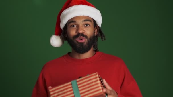 戴着圣诞圣诞圣诞礼帽的非洲裔年轻人被隔离在绿墙背景之外 手持礼品盒 — 图库视频影像
