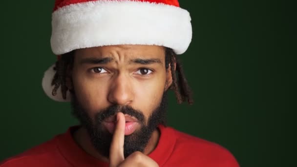 年轻快乐的非洲男人戴着圣诞圣塔帽 在绿墙的背景上表现出沉默的姿态 — 图库视频影像