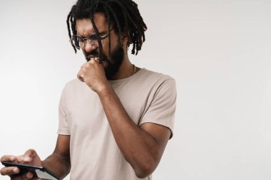 Sinirli, negatif Afrikalı bir adamın, gözlüklü, gri duvar arkaplanından izole edilmiş cep telefonuyla oyun oynaması.