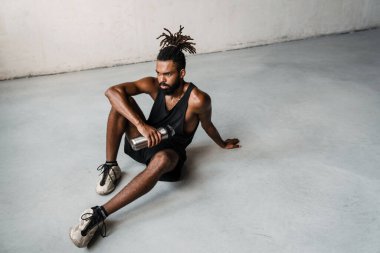Odaklanmış Afro-Amerikan sporcunun yerde oturmuş, su şişesiyle görüntüsü.