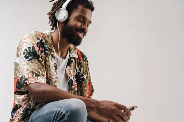ヘッドフォンで音楽を聴きながら携帯電話を使用して灰色の壁の背景に隔離された肯定的な陽気なアフリカ人の男のイメージ — ストック写真