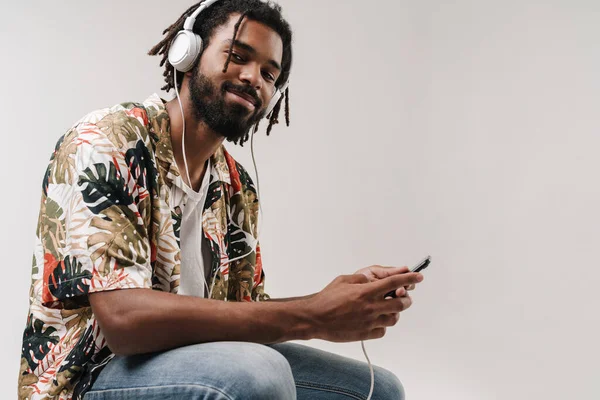 ヘッドフォンで音楽を聴きながら携帯電話を使用して灰色の壁の背景に隔離されたアフリカの男の写真 — ストック写真