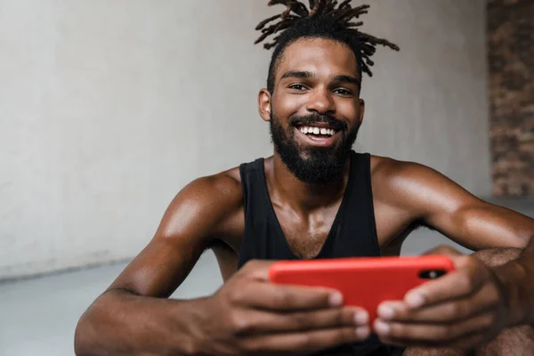 图为非洲裔美国运动员坐在室内用手机微笑的情景 — 图库照片