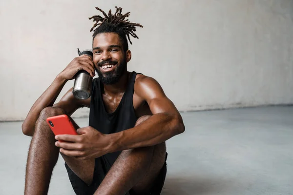 Obrázek Usmívajícího Afroamerického Sportovce Používajícího Mobilní Telefon Při Pití Vody — Stock fotografie