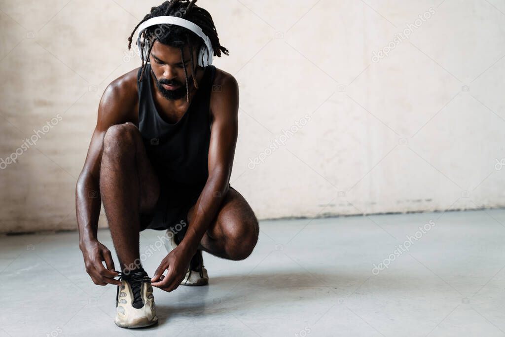 Image of african american sportsman in headphones tying up his shoelaces while working out indoors