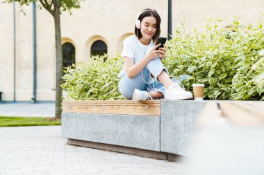 Mutlu, iyimser Asyalı bir kadının cep telefonu kullanırken ve kulaklıkla müzik dinlerken görüntüsü