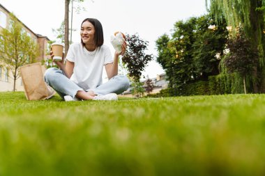 Mutlu genç Asyalı kadın tasviri çimlerin üzerinde oturmuş elinde kruvasan tutuyor ve kahve içiyor.