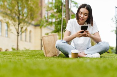 İyimser, genç Asyalı bir kadının cep telefonu kullanarak çimlerin üzerinde oturduğu fotoğraf.