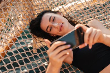 Sahilde hamakta yatarken cep telefonuyla selfie çeken genç bir kız görüntüsü.