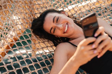 Sahilde hamakta yatarken cep telefonuyla selfie çeken genç bir kız görüntüsü.