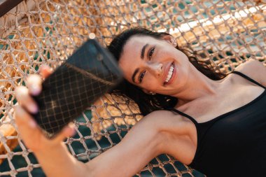Sahilde hamakta yatarken cep telefonuyla selfie çeken genç bir kız görüntüsü.
