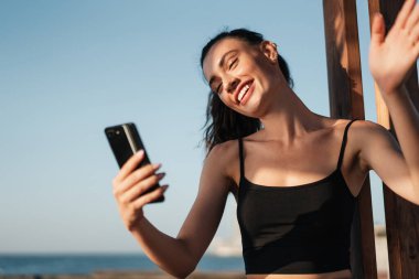 Genç, neşeli bir kızın el salladığı ve yaz plajında cep telefonuyla selfie çektiği bir resim.