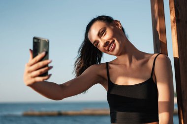 Yazın sahilde cep telefonuyla gülümseyen ve selfie çeken genç bir kız resmi.