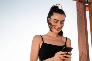 Sahilde cep telefonu kullanan mutlu, pozitif, genç bir fitness kadını görüntüsü.