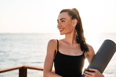 Sahilde elinde spor halısı tutan genç ve güzel bir jimnastik kadını hayal et.