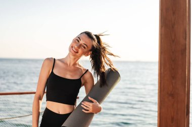 Sahilde elinde spor halısı tutan neşeli, genç ve güzel bir jimnastik kadının fotoğrafı.
