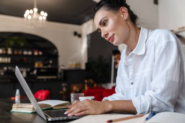 İçerideki kafede otururken dizüstü bilgisayarda çalışan gülümseyen genç kadın.