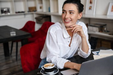 İçerideki kafede otururken dizüstü bilgisayarda çalışan gülümseyen genç kadın.