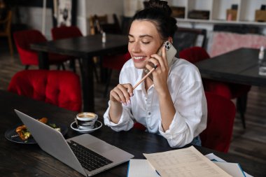 Laptop bilgisayarında çalışan gülümseyen genç bir kadın kafede oturmuş, cep telefonuyla konuşuyor.