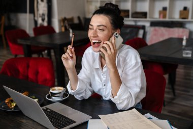 Laptop bilgisayarında çalışan gülümseyen genç bir kadın kafede oturmuş, cep telefonuyla konuşuyor.