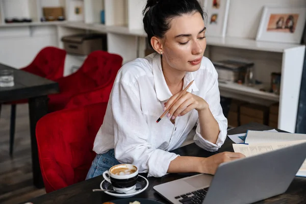 Güzel, kendine güvenen, ciddi, genç bir kadın kafenin içinde dizüstü bilgisayarla çalışıyor.