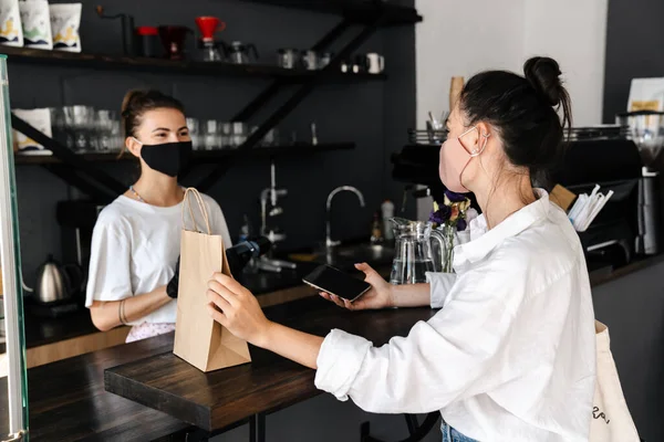Woman barista wearing medical mask giving takeaway order at the cafe counter to a client